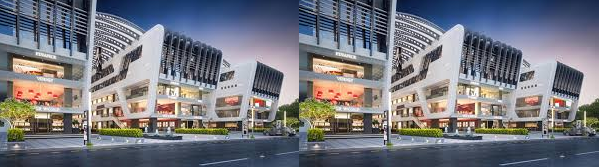 Modern commercial building with a sleek design and large windows, featuring illuminated storefronts and greenery in front, captured during twilight.